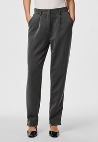 Grau gefaltete Hose aus einem strukturierten Stoff. Verfügt über einen Knopfverschluss, Seitentaschen und ein schmal zulaufendes Design. Schlichte schwarze Schuhe sichtbar.