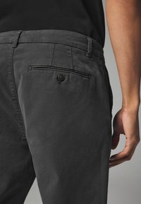 Graue Baumwollhose mit einer Gesäßtasche mit Knopfverschluss und einer glatten Textur. Der Bund ist flach mit Gürtelschlaufen.