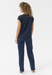 T-shirt bleu marine à manches courtes avec des détails en dentelle sur les épaules, dos en découpe en trou de serrure, associé à un jean droit bleu foncé et des escarpins clairs.