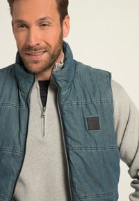 Jeansstil blå väst med hög krage, dragkedja och sömnadsdetaljer. Bärs över en ljusgrå zip-up tröja. Varumärkeslogotyp på västen.