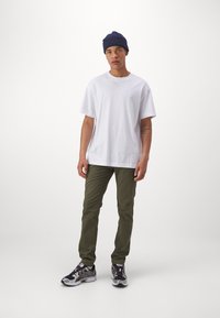 White cotton t-shirt, olive green slim-fit pants, and black athletic sneakers with gray accents. Navy knit beanie on head.