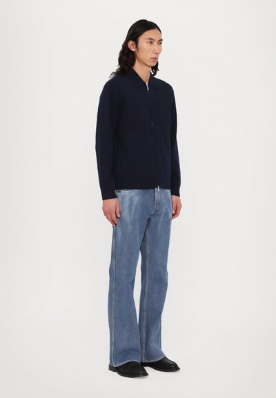 Cardigã azul-marinho com fecho éclair e gola. Combinado com jeans azuis claros em corte flare. Sapatos pretos de ponta redonda completam o conjunto.