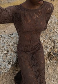 Hnedé čipkované šaty s dlhými rukávmi, zdobené zložitými kvetinovými vzormi a priliehavým strihom, na pozadí skalnatého terénu.