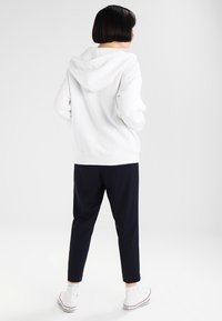Sweat-shirt à capuche blanc avec des poignets côtelés, associé à un pantalon tapered bleu marine. Le look est complété par des baskets blanches avec des accents rouges.