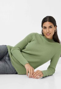 Pull à manches longues côtelé de couleur vert clair, avec un col haut et un tissu texturé. Porté avec un jean gris, affichant une coupe décontractée.