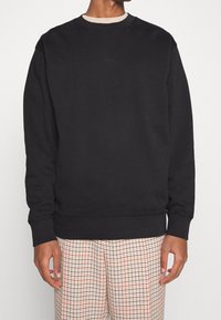 Svart sweatshirt med rund hals, ribbad mudd och nederkant, med en diskret logotyp på bröstet, matchad med mönstrade beige byxor.