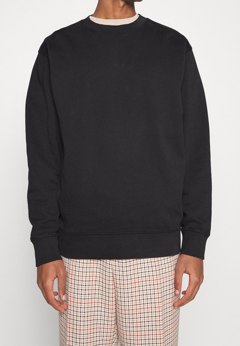 Svart sweatshirt med rund hals, ribbad mudd och nederkant, med en diskret logotyp på bröstet, matchad med mönstrade beige byxor.