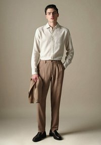 Helles beige Button-Up-Hemd, braune maßgeschneiderte Hose und schwarze Loafers. Das Model hält einen braunen Pullover und steht vor einem neutralen Hintergrund.