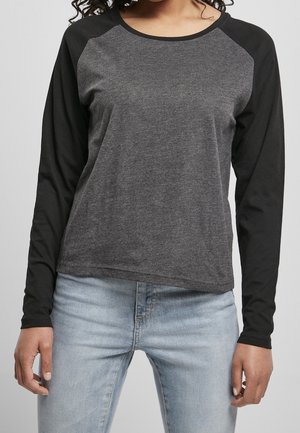 Person trägt ein dunkelgraues Langarmshirt mit schwarzen Ärmeln und hellblauen Jeans und steht vor einfachem Hintergrund.