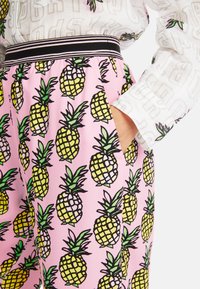 Rosa gemusterte Hosen mit gelben Ananas und schwarzen Umrandungen, elastischer Bund mit schwarz-weißen Streifen, funktionale Taschen.