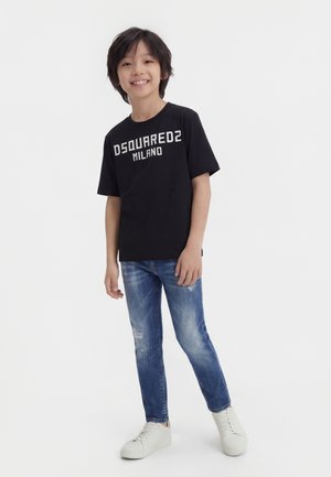 Ragazzo sorridente che indossa una t-shirt nera DSQUARED2 Milano, jeans blu strappati e sneakers bianche, in piedi su uno sfondo bianco.