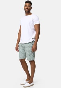 T-shirt en coton blanc, shorts verts à motifs en losanges, espadrilles blanches. Le modèle se tient avec confiance, les bras détendus sur les côtés.