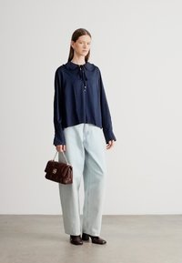 Μπλούζα σε ναυτικό μπλε με μακριά μανίκια και γιακά, συνδυασμένη με φαρδιά ανοιχτό μπλε παντελόνια. Αξεσουάρ: σκούρο καφέ τσάντα. Λείο ύφασμα.