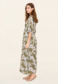 Grünes, blumiges Maxikleid mit kurzen Ärmeln, das mit blau-weißen Blattmustern verziert ist. Kombiniert mit hellen, flachen Schuhen.