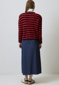 Crvena i crna prugasta pletena dukserica s rebrastom teksturom, nošena preko traper suknje. Outfit uključuje cipele neutralnih tonova i bijelu kragnu.