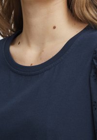 T-shirt en coton bleu marine avec un col rond, présentant une texture subtile et des accents œillets sur les manches. Tissu lisse avec peu de détails.