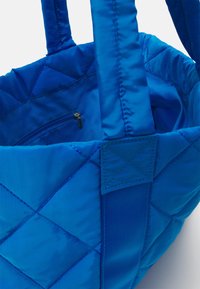 Vadderad blå tote-väska med slät satinfinish, med två stabila handtag och en inre ficka med dragkedja. Strukturerat diamantmönster på utsidan.