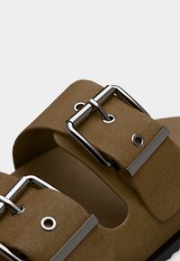 Κοντινό πλάνο από λουράκια καφέ σουέντ σανδαλιών με ασημί μεταλλικά αγκράφα και οπές σε λευκό φόντο.