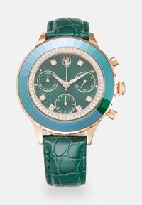 Orologio rotondo con cinturino in pelle verde texturizzata. Il quadrante presenta uno sfondo verde scuro, dettagli in cristallo e tre sub-quadranti. Hardware color oro.
