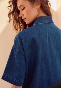 Camisa de mezclilla en azul oscuro, con cuello, mangas cortas y detalles de costura visibles, presentada con un acabado texturizado.