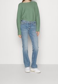 Vaaleanvihreä neule, joka on väljät mitoitukset, yhdistettyinä haalistuneisiin sinisiin suoriin housuihin ja valkoisiin tennareihin, joissa on ruskeita yksityiskohtia. Minimalistinen muotoilu, ilman kuvioita.