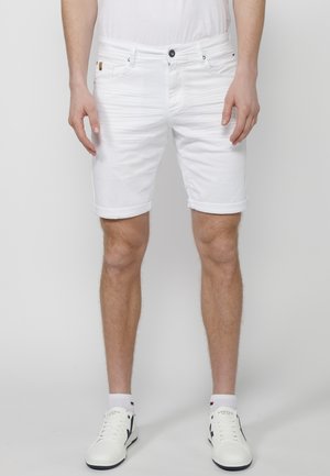 Shorts de mezclilla blanca con dobladillos enrollados, cinco bolsillos y cierre de botón negro. Combinados con zapatillas blancas y calcetines hasta el tobillo.