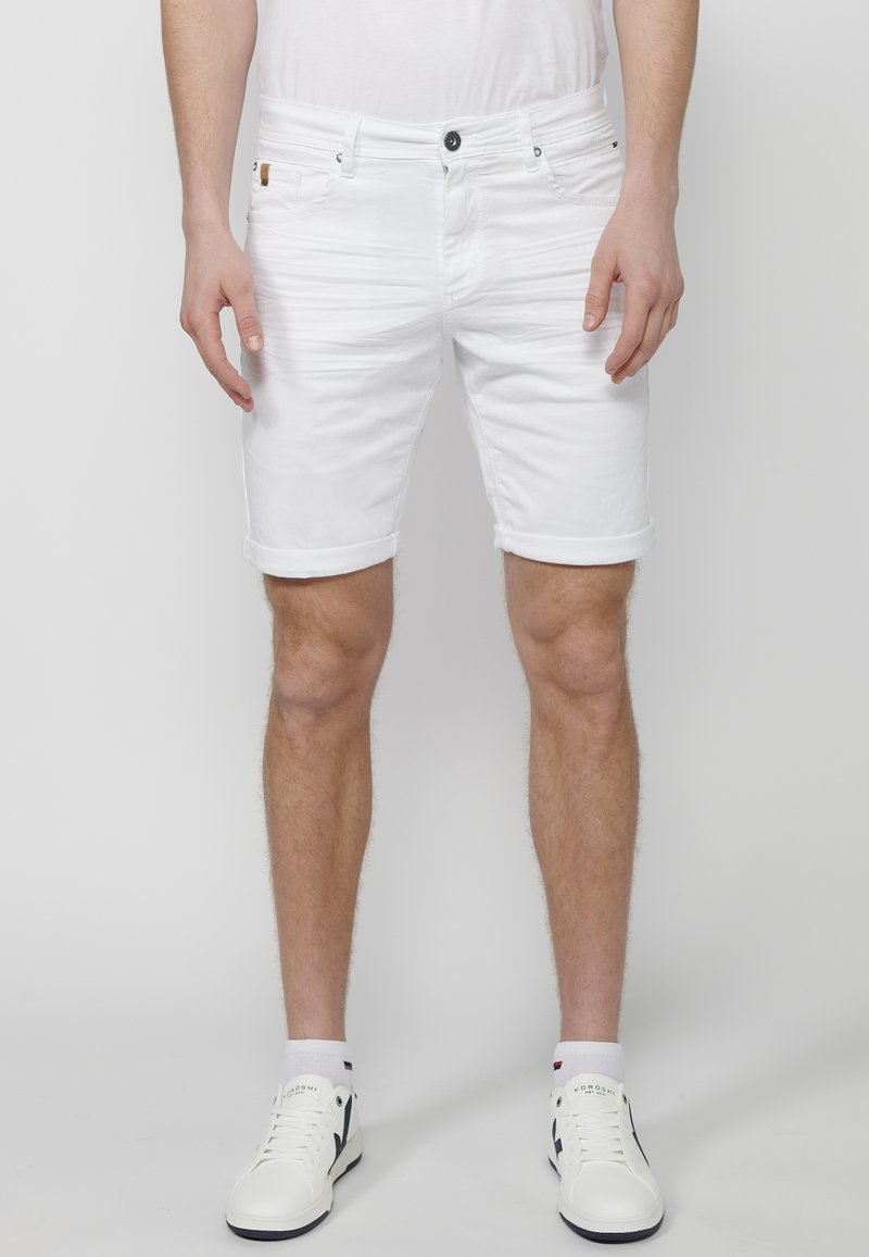Shorts de mezclilla blanca con dobladillos enrollados, cinco bolsillos y cierre de botón negro. Combinados con zapatillas blancas y calcetines hasta el tobillo.