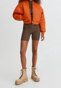 Veste bomber matelassée orange, body marron côtelé et bottines de combat beiges à lacets. Matériaux texturés avec des couleurs et des motifs contrastés.