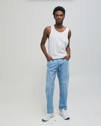 Nuori mies seisoo käsineet taskuissa, yllään valkoinen hihaton paita, vaaleansiniset farkut ja valkoiset tennarit yksinkertaisen taustan edessä.