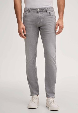 Mężczyzna noszący dopasowane jasnoszare dżinsy i białe trampki, stojący na tle jasnego, jednolitego tła.