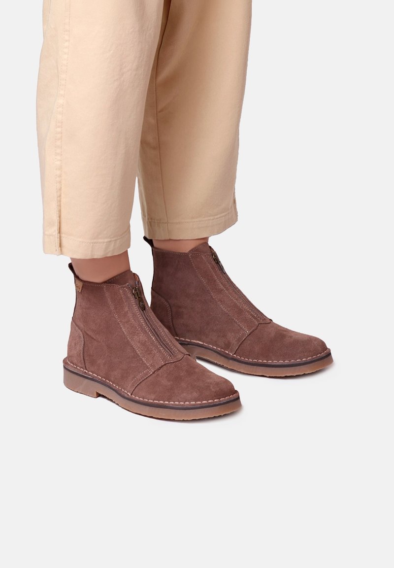 Bottines en daim marron avec fermetures éclair à l'avant, portées avec un pantalon beige court sur un fond clair.