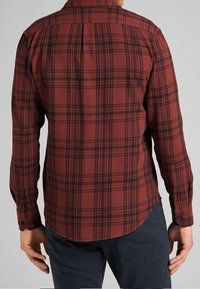 Camisa de cuadros rojos con detalles en negro, de botones, de mangas largas y con dobladillo curvado. Tejido suave con un acabado texturizado, que presenta costuras visibles.