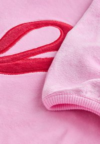 Sweat-shirt rose avec une texture douce et pelucheuse, orné d'un motif ondulé rouge. Le poignet est fini avec un bord-côte.