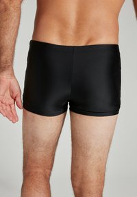 Schwarze Badeshorts aus dehnbarem Stoff, mit einer taillierten Passform, die bis zur Mitte des Oberschenkels reicht, und einer glatten Textur sowie minimalen Nähten.