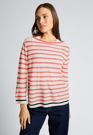 Donna che indossa un maglione bianco a maniche lunghe con strisce orizzontali corallo e jeans scuri, in piedi contro uno sfondo chiaro e uniforme.