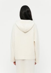 Maglione con cappuccio color crema realizzato in un tessuto morbido e strutturato, con spalle scese e orlo a coste. Mostra la vista posteriore.