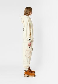 Persona vestida con sudadera oversized de color crema y pantalones cargo con un gran bolsillo, acompañada de botas gruesas marrones, de pie de perfil.