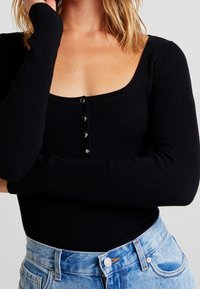 Femme portant un haut noir à manches longues côtelé avec quatre boutons et un jean en denim bleu clair taille haute, les bras croisés.