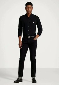 Νεαρός άνδρας ντυμένος με μαύρο πουκάμισο, μαύρο παντελόνι, μοτίβο γκρι κάλτσες, μαύρες loafers και ασημένιο ρολόι χειρός.