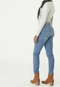 Hochtaillierte blaue Jeans mit schlanker Passform, ausgestattet mit Gesäßtaschen, kombiniert mit braunen Wildleder-Stiefeletten auf einem strukturierten beigen Hintergrund.