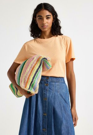 Kvinde med krøllet mørkt hår iført ferskenfarvet t-shirt og knappet denimnederdel, der holder en stor farverig stribet strikketaske under armen.