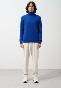 Niebieski sweter w golfie z miękkiej tkaniny, połączony z jasnobeżowymi spodniami w prążki. Model nosi białe trampki i stoi na neutralnym tle.