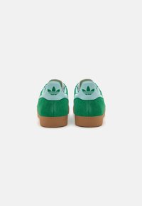 Tenis de camurça verdes com detalhes em azul claro, apresentando uma sola de borracha texturizada e um logotipo no calcanhar. Design de cano baixo.