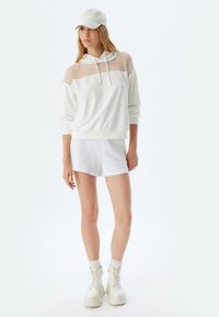 Felpa bianca con inserti in rete sulle spalle, abbinata a pantaloni corti bianchi e stivali alla caviglia bianchi chunky. Tessuto morbido, design casual.