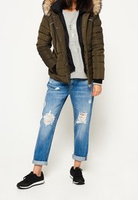 Chaqueta acolchada verde oliva con capucha forrada de piel, combinada con una sudadera gris y vaqueros desgastados azul claro, completada con zapatillas negras.