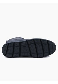 Suela de zapatilla de goma negra con patrones de relieve profundos, que presenta un logo rectangular en el centro y bordes reforzados para mayor durabilidad.