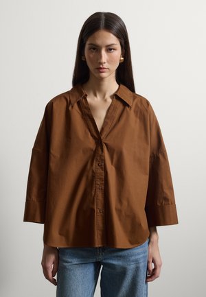 Mujer de cabello largo y oscuro, con pendientes dorados, camisa marrón suelta con botones y mangas anchas, y vaqueros azules, de pie frente a un fondo liso.