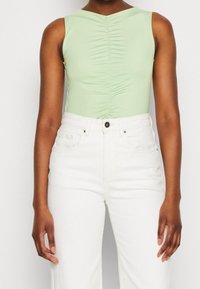 Ljusgrön ärmlös bodysuit med ruching-detalj framtill, parat med högmidjade vita byxor; slät textur och figurnära design.