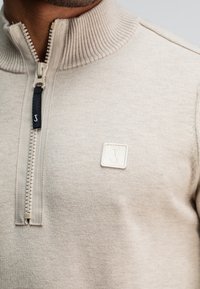 Beige zip-up trui met een geribde kraag, voorzien van een label met een subtiel logo. Glad textuur en een eenvoudig ontwerp.
