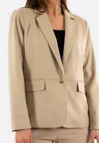 Blazer beige con un diseño ajustado, cierre de botón único y dos bolsillos frontales. Textura suave y hombros estructurados.